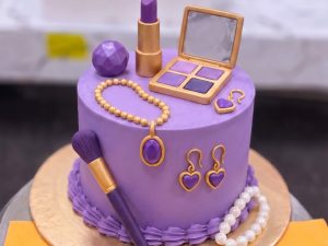 Purple Doll Glam Cake in Lahore — A deep purple cake with an eyeshadow palette, lipstick, earrings and a pearl necklace laid across the top and