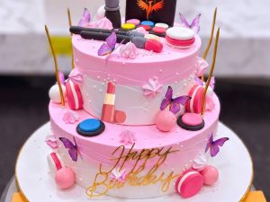 Two Tier Barbie Makeup Cake in Lahore — A two tier pink makeup cake — a white lower tier with a piped name, a pink upper tier with butterflies, lipsti
