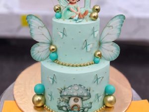 Fairy Doll Cake in Lahore — A mint green two tier cake with a fairy doll perched on top, gold wings, dragonflies on wires and gold spheres