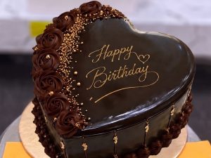 Chocolate Birthday Heart Cake in Lahore — A glossy dark chocolate heart cake with gold "Happy Birthday" lettering, a piped rose border and gold sprinkle