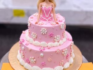 Barbie Bow Doll Cake in Lahore — A two tier pink doll cake with a Barbie in a bow headband, white blossoms across both tiers and neat shell pip