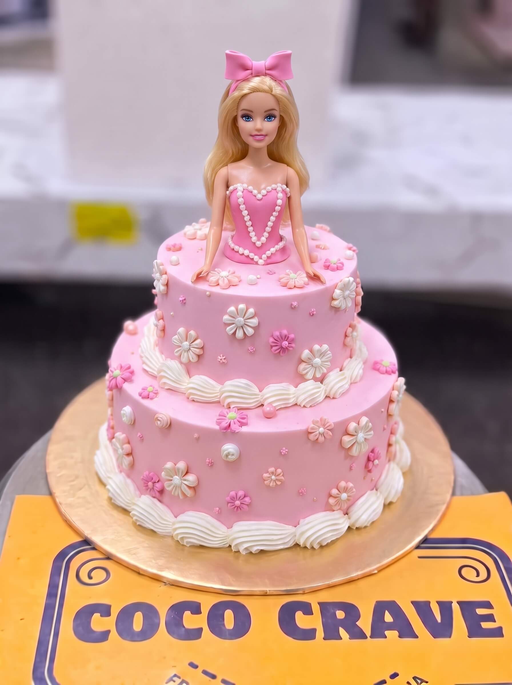 Barbie Bow Doll Cake in Lahore — Coco Crave Lahore Barbie Bow Doll Cake in Lahore — A two tier pink doll cake with a Barbie in a bow headband, white blossoms across both tiers and neat shell pip